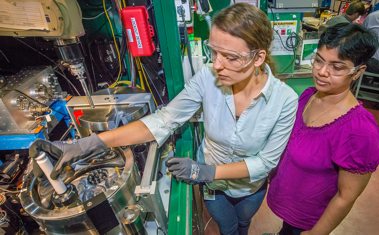 At left, a researcher wearing a light-blue button-down shirt and blue jeans, protective eyewear, and gray gloves loads a small white cylindrical sample holder into a silver-toned metal dewar framed by dark-green metal. At right, another researcher wearing a bright purple blouse, dark pants, and protective eyewear, stands by to observe.