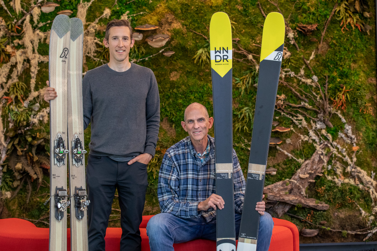 Two people in front of a red couch holding skis.