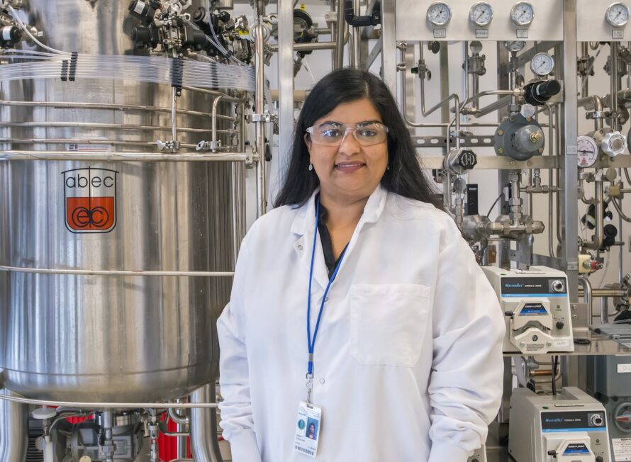 A woman in a white lab coat and safety glasses stands in front of a silver-toned bioreactor: a large vat with many connected tubes, knobs, sensors, and other machinery.