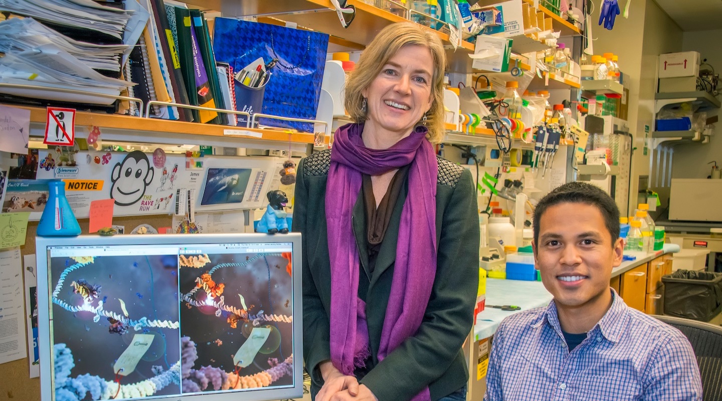 A woman with a purple scarf and a man in a blue checked shirt sit at a desk with many containers and books on shelves in the background. Next to them is a computer monitor showing a 3D rendering of several molecules.