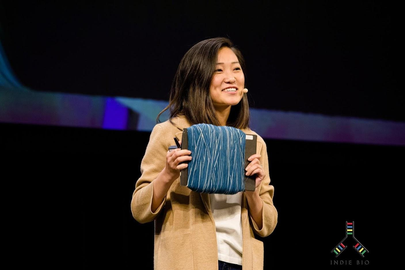 A person with long dark hair wearing a yellow blazer and a headset holding denim yarn.