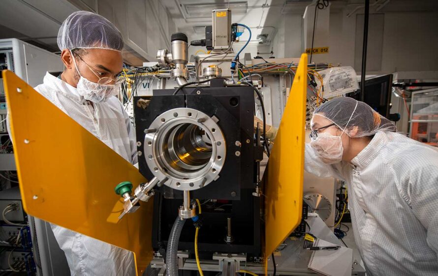 Two researchers donning personal protective equipment inspect a mirror chamber used to create custom and dynamic illumination patterns for testing the limits of semiconductor materials.