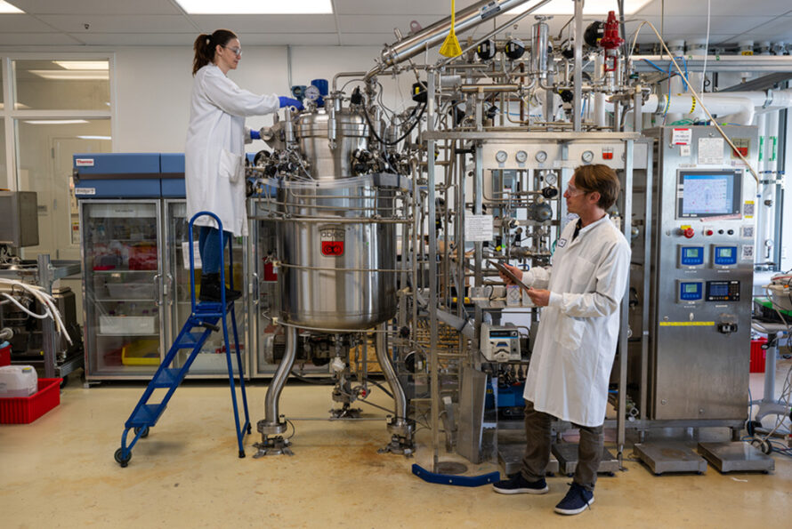 Two researchers wearing white lab coats and safety goggles working with large machinery in the Lab.