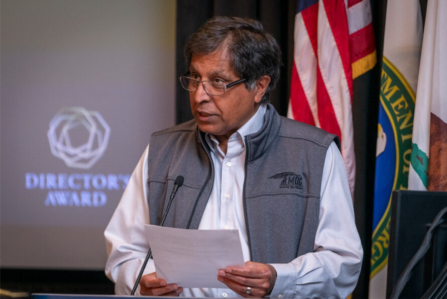 Sudip Dosanjh, wearing glasses and a grey vest, reads a sheaf of papers from a lectern