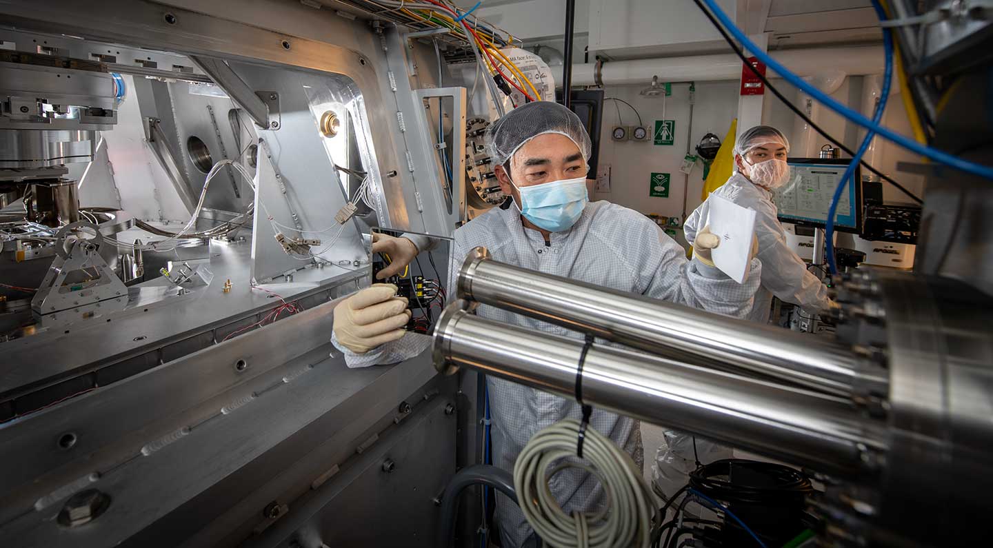 Two researchers wearing white lab coats in a lab with large metal machinery and blue cables.