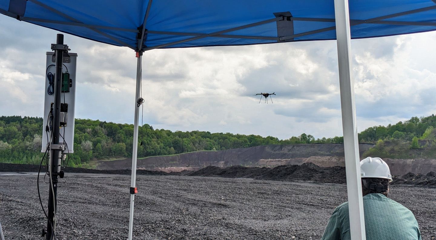 Man sits beneath tent cover while operating a drone flying above in the distance.