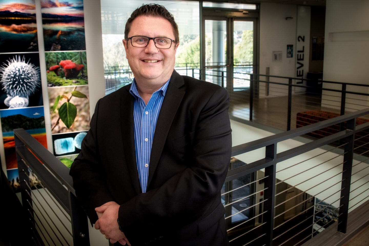 A man with glasses in black suit poses in front of close up pictures of plants