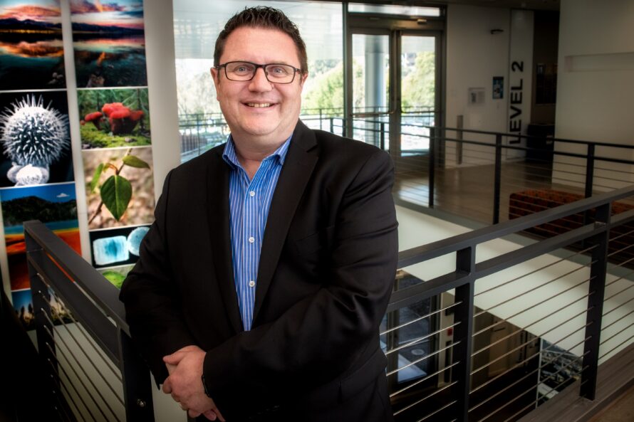 A man with glasses in black suit poses in front of close up pictures of plants