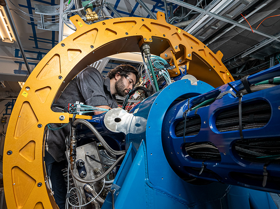 A person looks at something hidden behind GRETA's cylindrical blue metal structure, which holds numerous cables. The person is framed by a large, circular yellow arch that connects to the blue assembly.