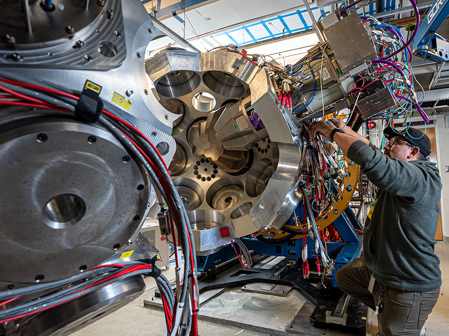 A person touches the outside of half of a large metal sphere. The central structure has a circular array of large, machined openings and visible internal components, with cables running along the outside.