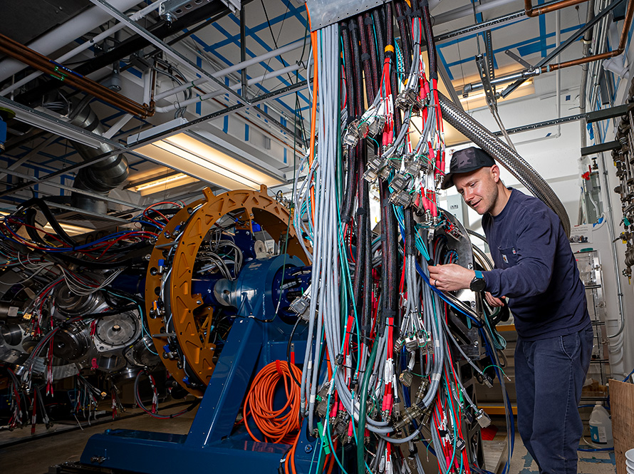 A person holds one of dozens of cables neatly draped from a tray several feet in the air. GRETA's frame, fully instrumented with detectors and cables, is visible in the background.