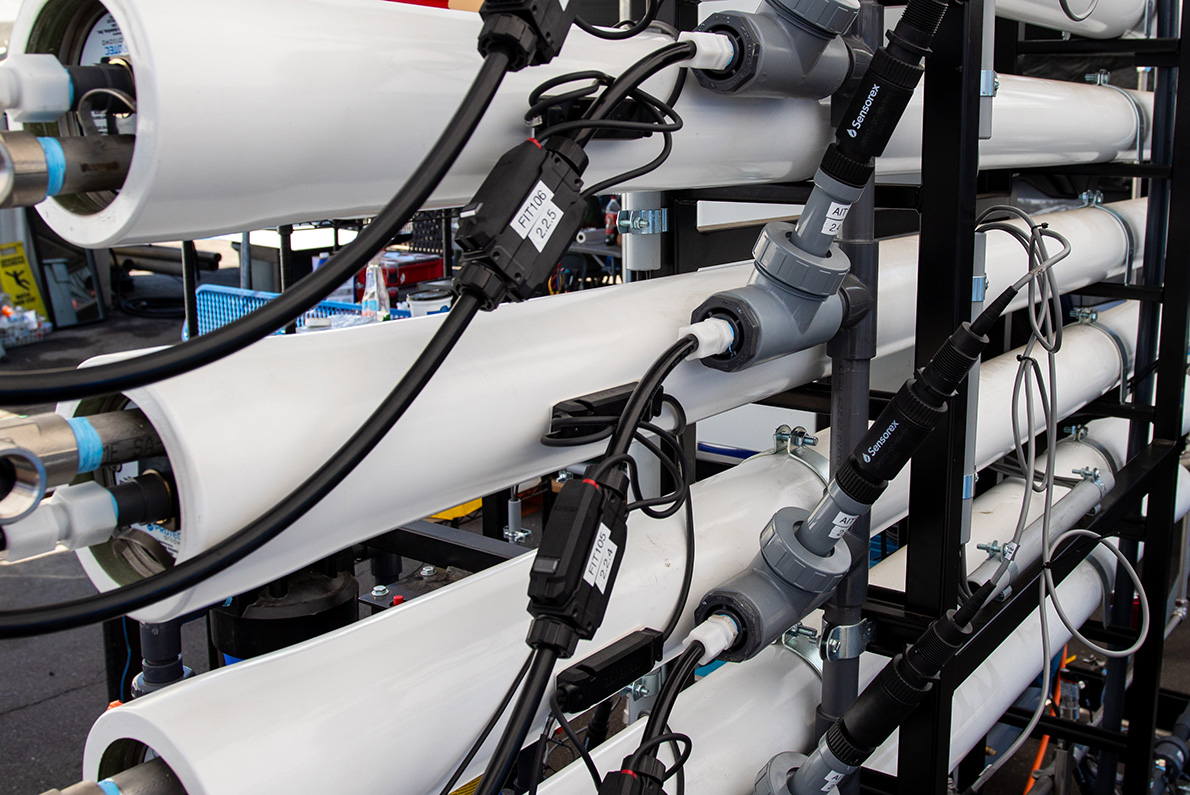 Industrial membrane technology setup for advanced osmosis, with interconnected white pipes, black metal frame, and attached electrical cords and wires