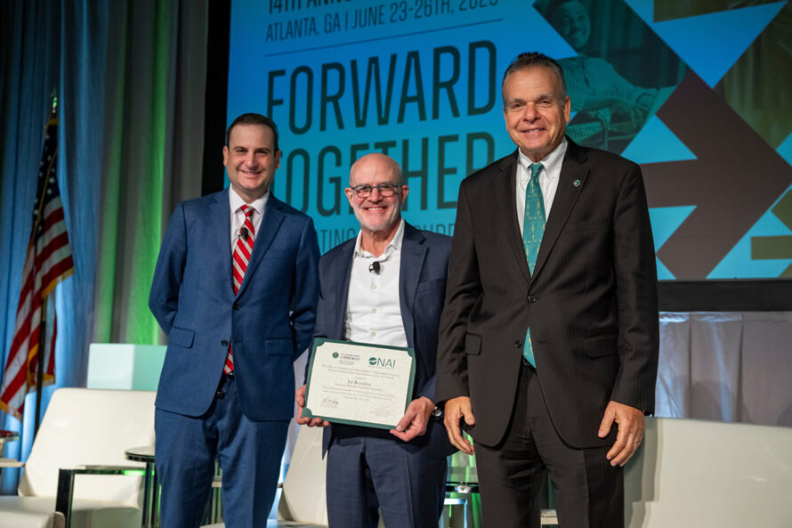 Three people in business suits stand together on a stage at an award ceremony, smiling at the camera. The person in the center holds a certificate.