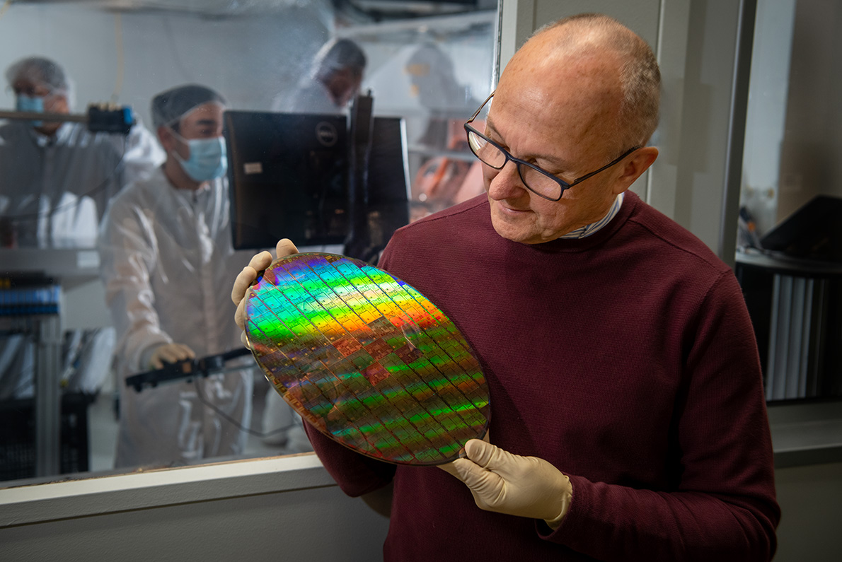 A man wearing a burgundy sweater, light-blue button-down shirt, and dark-rimmed eyeglasses stands in front of machinery made of transparent and black-toned materials while holding a large, rainbow-colored reflective disc in a clean room setting with white walls.