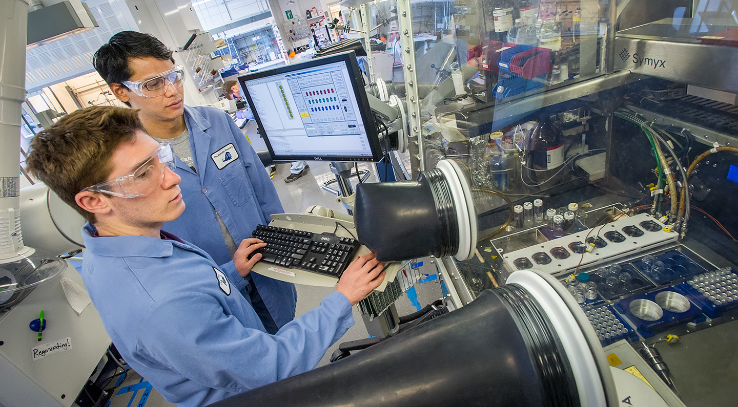 Two people in lab coats and safety goggles work at a computer and machine.
