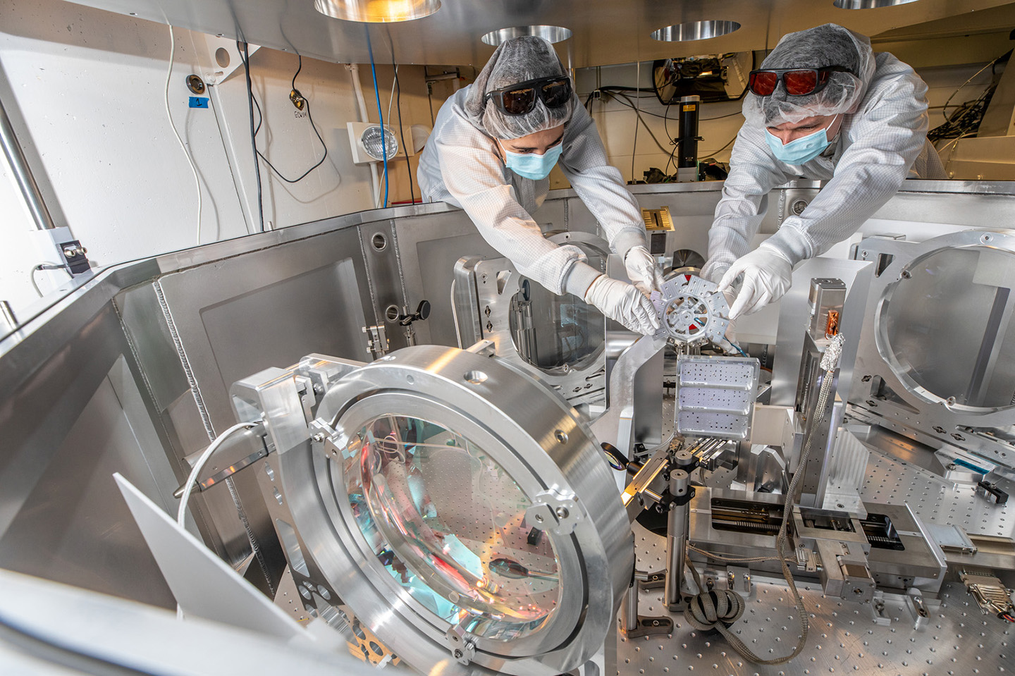 Two people in cleanroom suits install a small silver device in a small enclosure with lenses and other equipment.