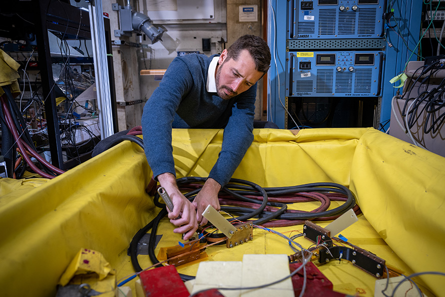 A person uses a wrench inside a yellow containment area in a laboratory filled with equipment and cables.