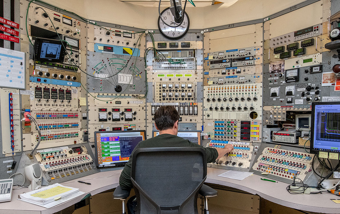 A person adjusts a setting at a console with multiple panels of knobs, switches, and screens.