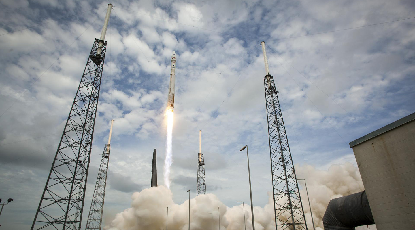 A rocket fires up into scattered clouds, causing clouds of dust on the ground.
