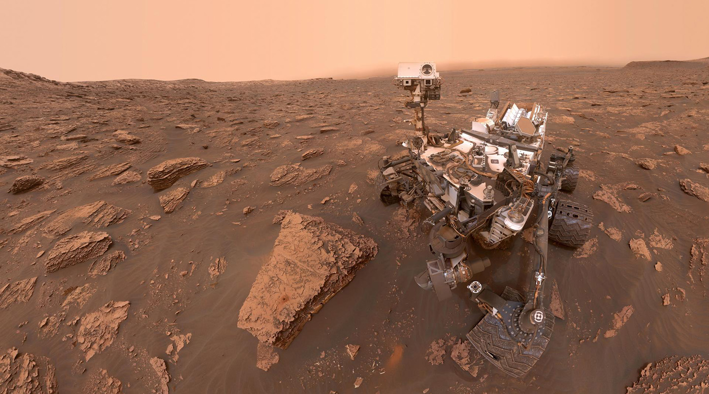 NASA's Curiosity rover surrounded by rocks and dust under a hazy orange sky, capturing a panoramic view of the Martian landscape with its onboard camera.