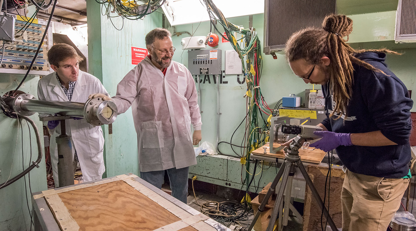 Three people standing in a lab space. One adjusts a piece of equipment on a tripod.