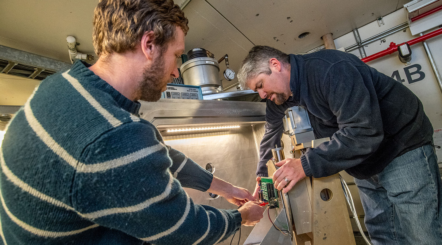 Two people work on electronics in a narrow enclosure.