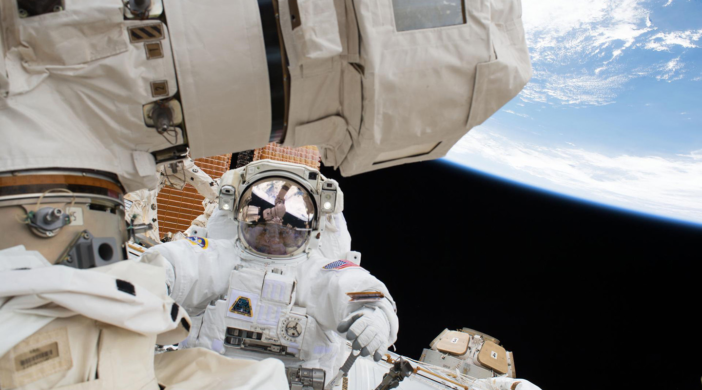 Person in a white spacesuit with a reflective face shield moves along the exterior of a section of the ISS. Earth is visible in the background.