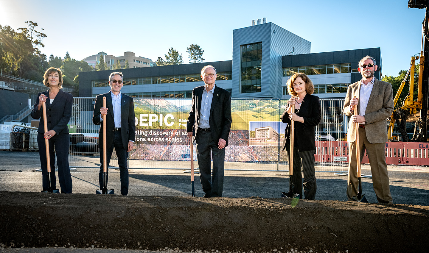 Five individuals stand with shovels at a groundbreaking ceremony in front of a large, new building.