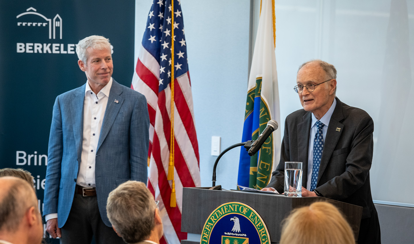 Two people dressed in suits stand near a podium facing a group of people in the foreground. The American flag is visible, along with the Berkeley Lab logo and the DOE logo.
