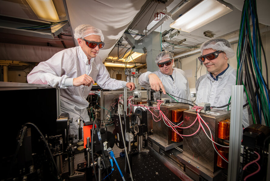 Three researchers in lab coats and hairnets working on a complex machine with wires and equipment, wearing protective orange-tinted glasses.