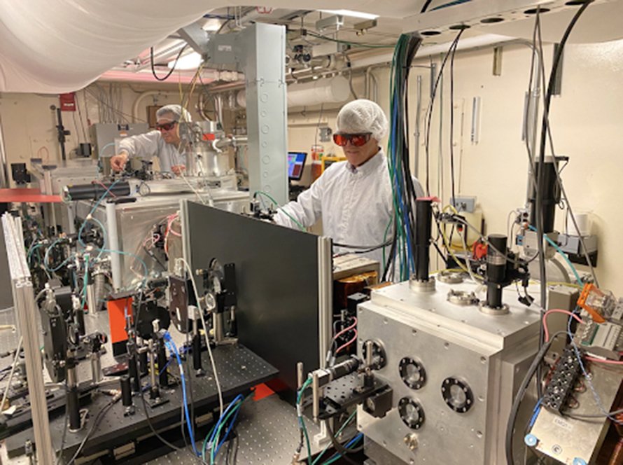 Two researchers in lab coats and hairnets working on a complex machine with wires and equipment, wearing protective orange-tinted glasses, with a large metal box and various instruments in the foreground.