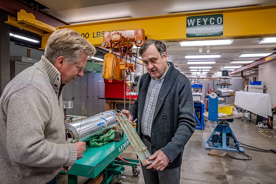 Two researchers speaking in a full laboratory. One researcher is holding a high-temperature superconductor that is roughly the size of a large ruler while the other researcher looks toward it.