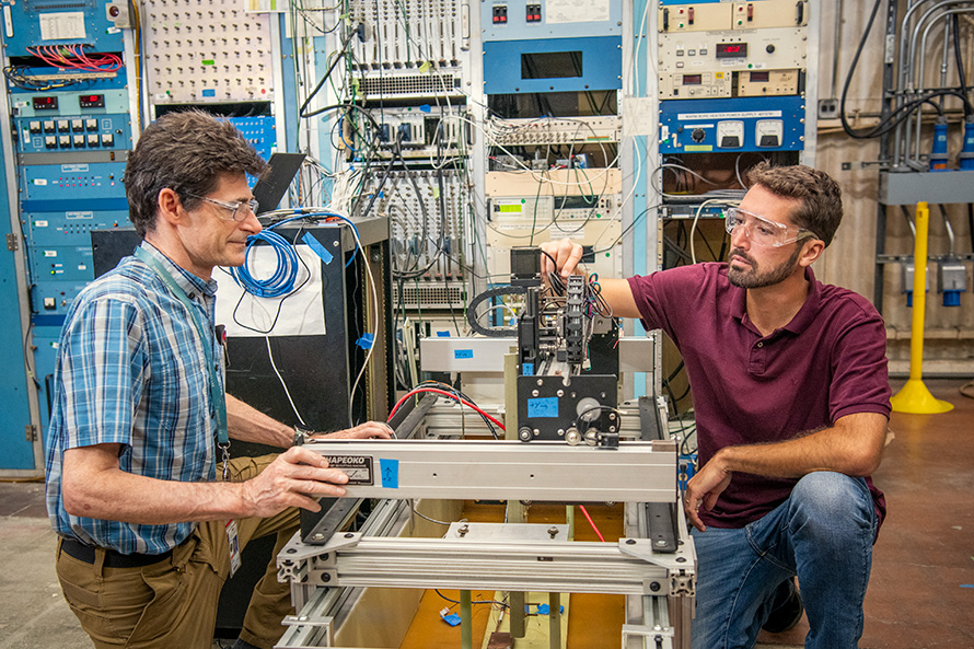 Two researchers inspect a high-temperature superconducting joint with a cryoscanner. A wall full of multicolored buttons, wires, and screens is in the background.