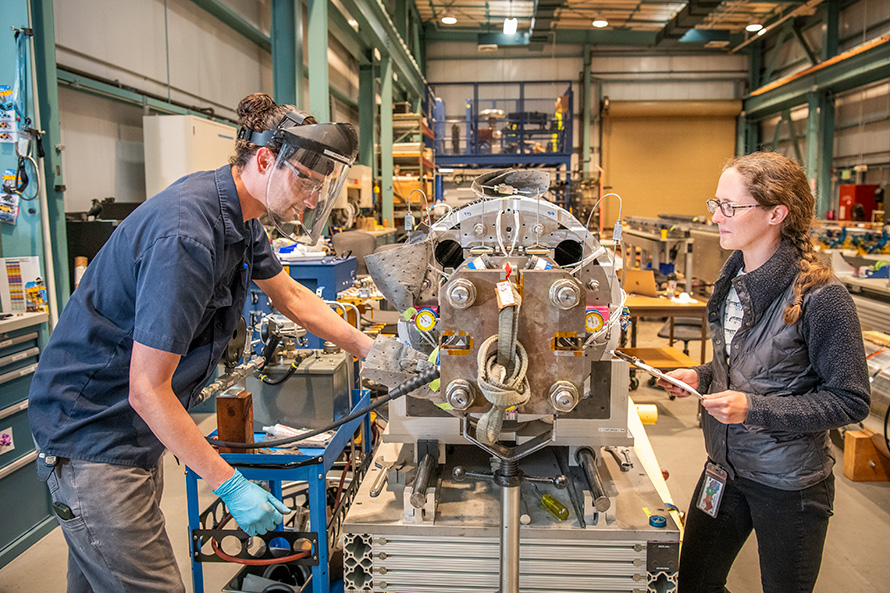Two researchers in a large machine shop working on either side of a grey quadrupole magnet.