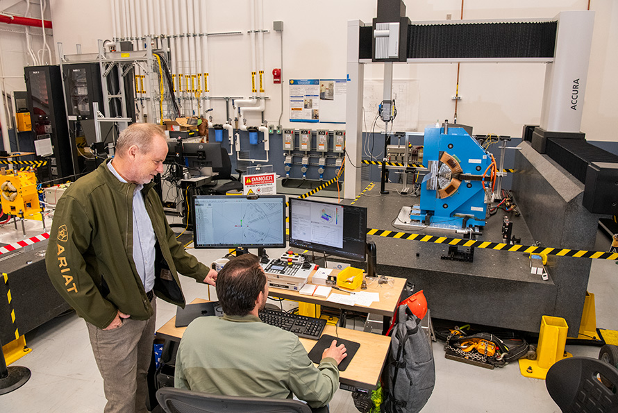 Two researchers sitting behind a computer desk in a large experimental facility. They are looking toward an electromagnet in the background.