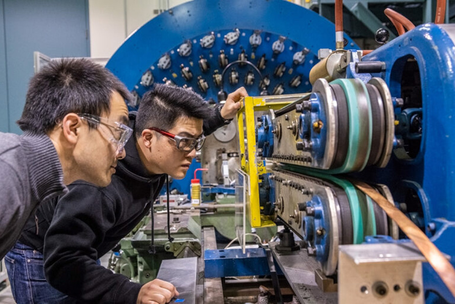 Two researchers in protective glasses inspect a copper colored magnet cable that is moving through a blue and yellow system.