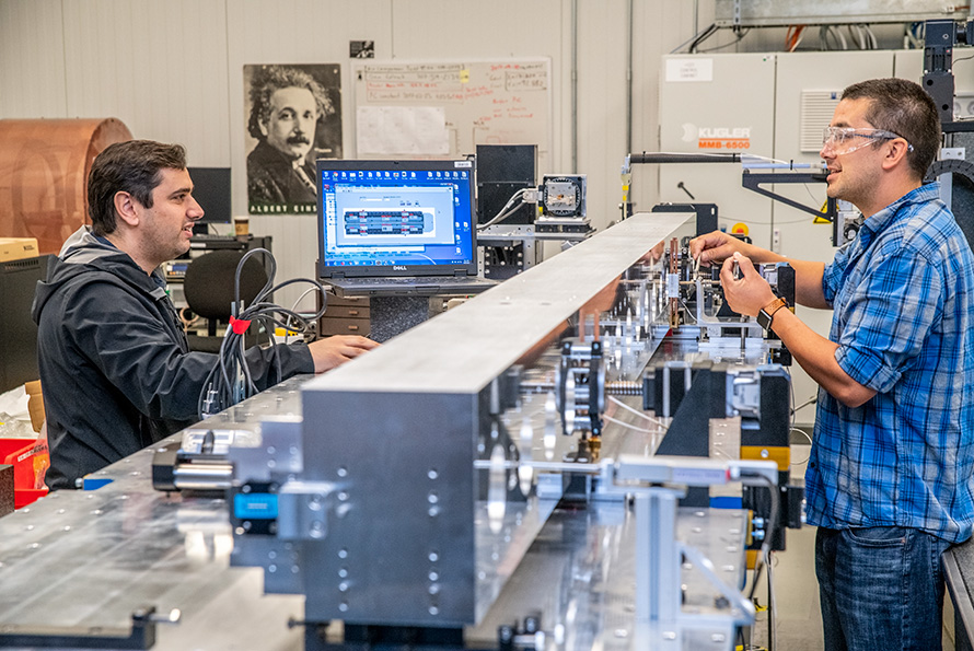 Two researchers talk across a long metal undulator while making adjustments.
