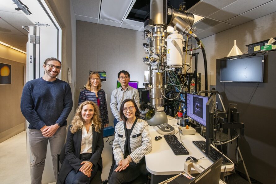 A group of five people are posed near a tall metallic instrument with multiple pipes and wires emerging from it. The instrument sits atop a desk with multiple monitors.