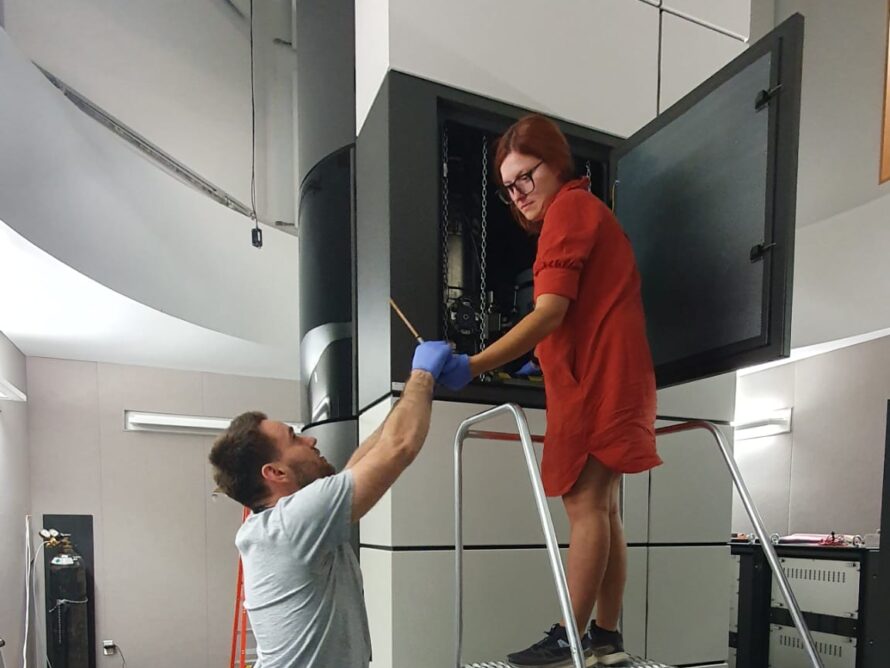 A woman stands on a step ladder in front of a floor-to-ceiling instrument within a column with a light and dark gray covering. A dark gray door the size of an oven door is open at the mid-room level, she and a male colleague are making adjustments to the components inside.
