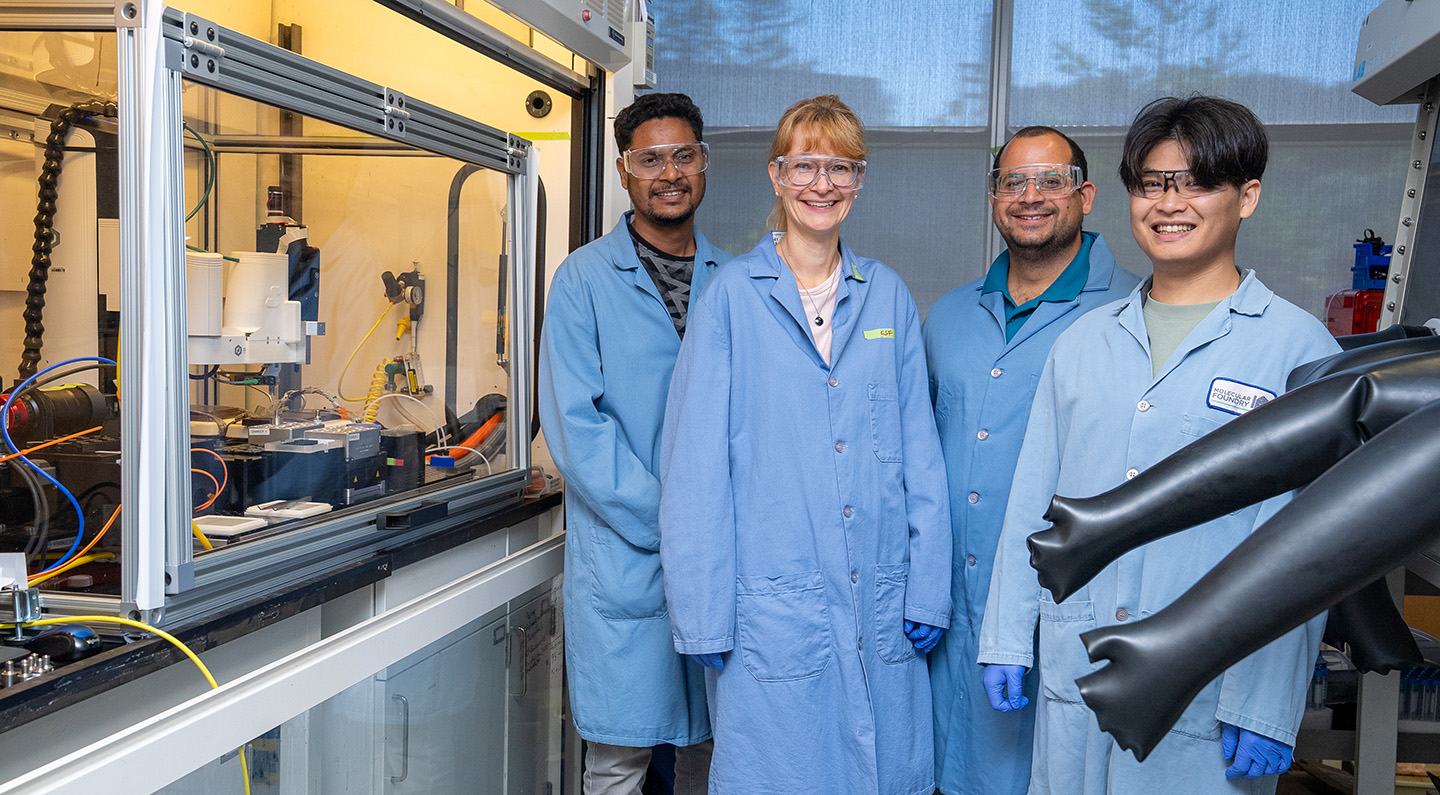 Four scientists in blue lab coats stand next to robot setup.
