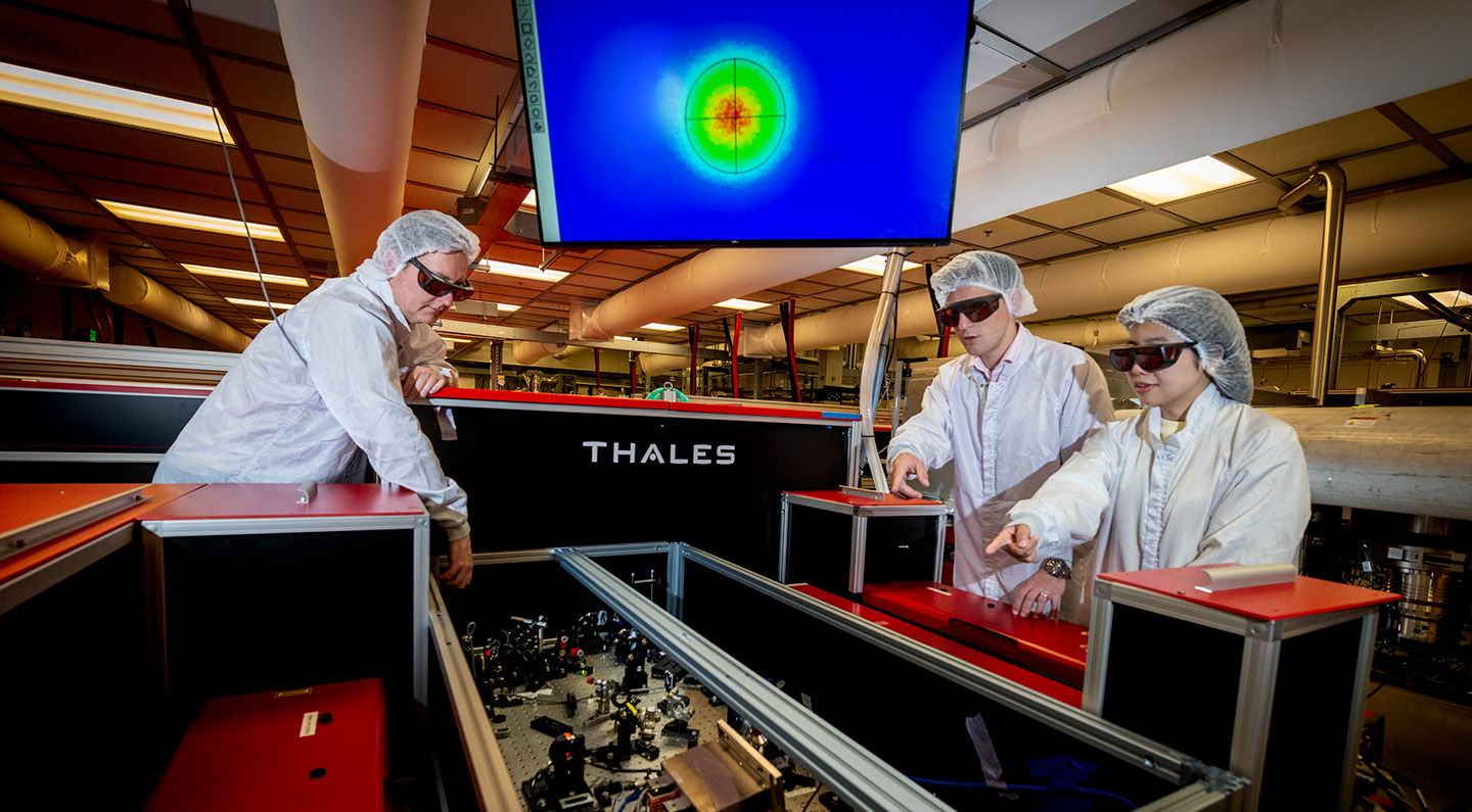 Three researchers in white coveralls align precision optics on the Berkeley Lab Laser Accelerator Petawatt laser. A monitor with colorful figures is in the background.