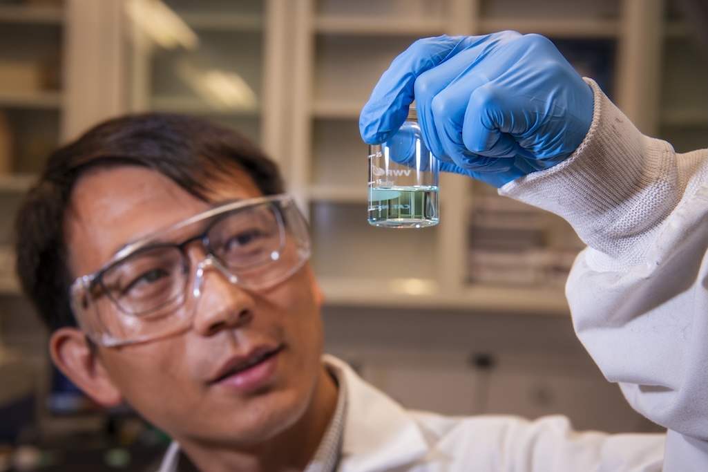 A close-up photograph of a man wearing glasses, a labcoat, and gloves who is holding up a small clear vessel containing light blue liquid.