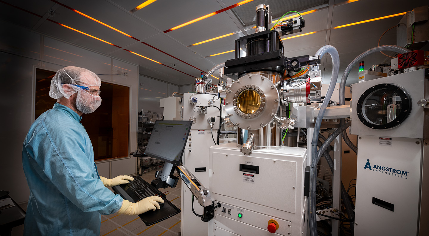 A researcher in light blue personal protective equipment stands at a computer stand which is connected to the cluster tool.