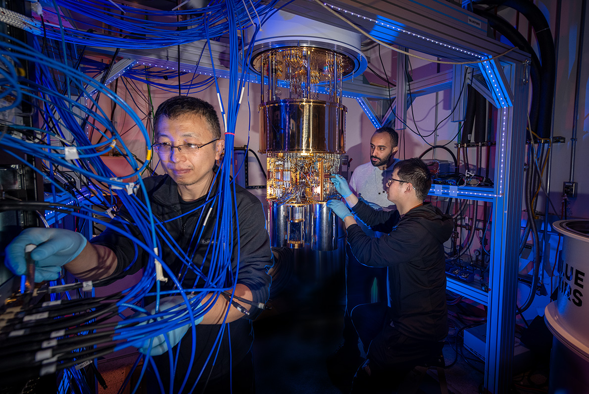 Three researchers work in a small lab space full of blue wires and a golden dilution fridge.