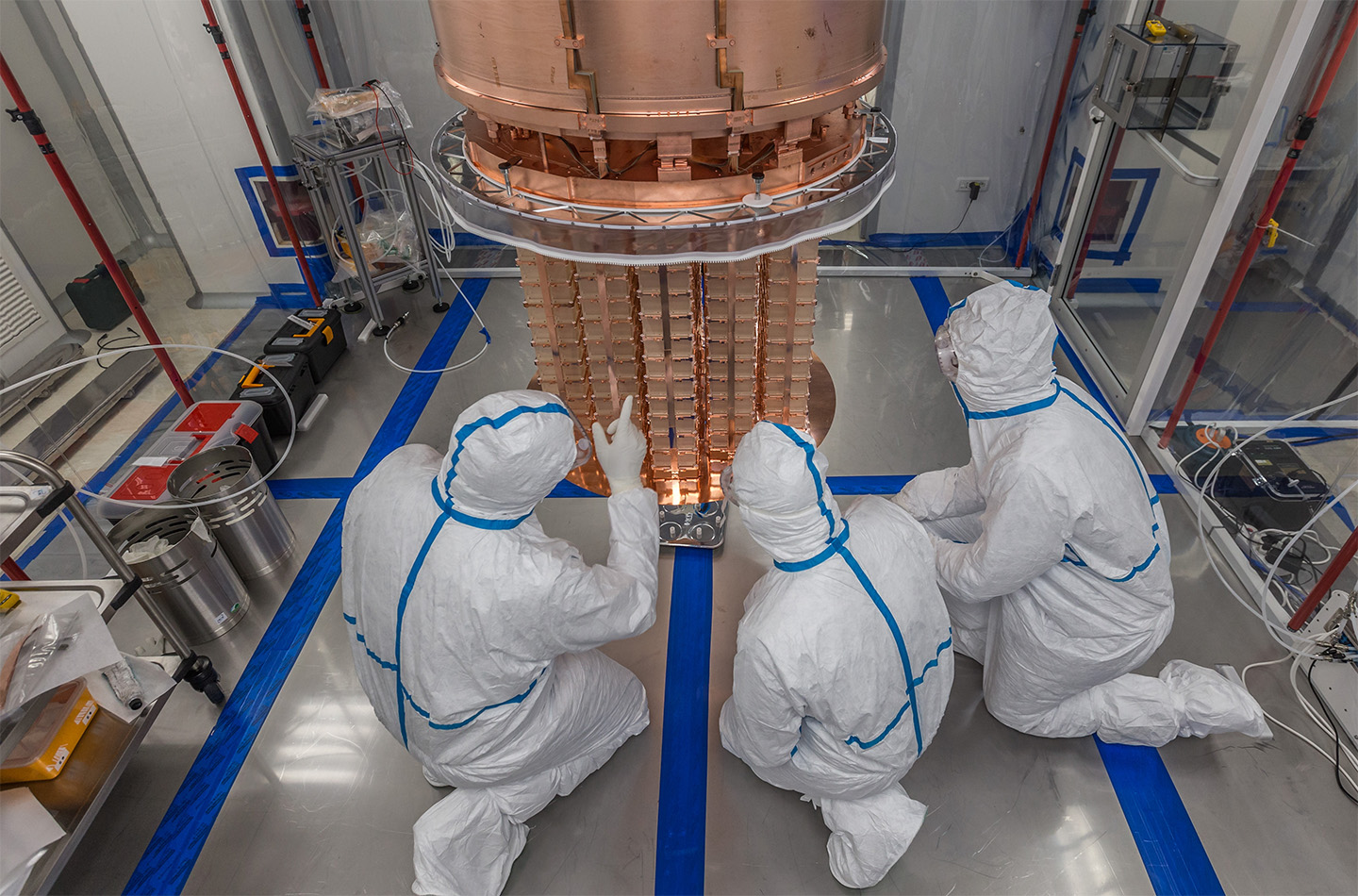 Three researchers in full cleanroom suits kneel around the CUORE experiment, working on a large suspended copper structure.