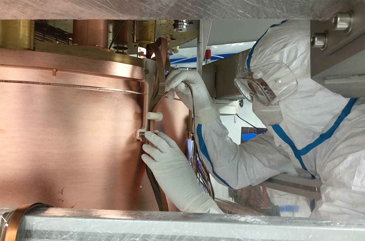 A person in full cleanroom attire wears a white protective suit, gloves, and safety glasses. They are attaching components to a large, cylindrical copper structure.