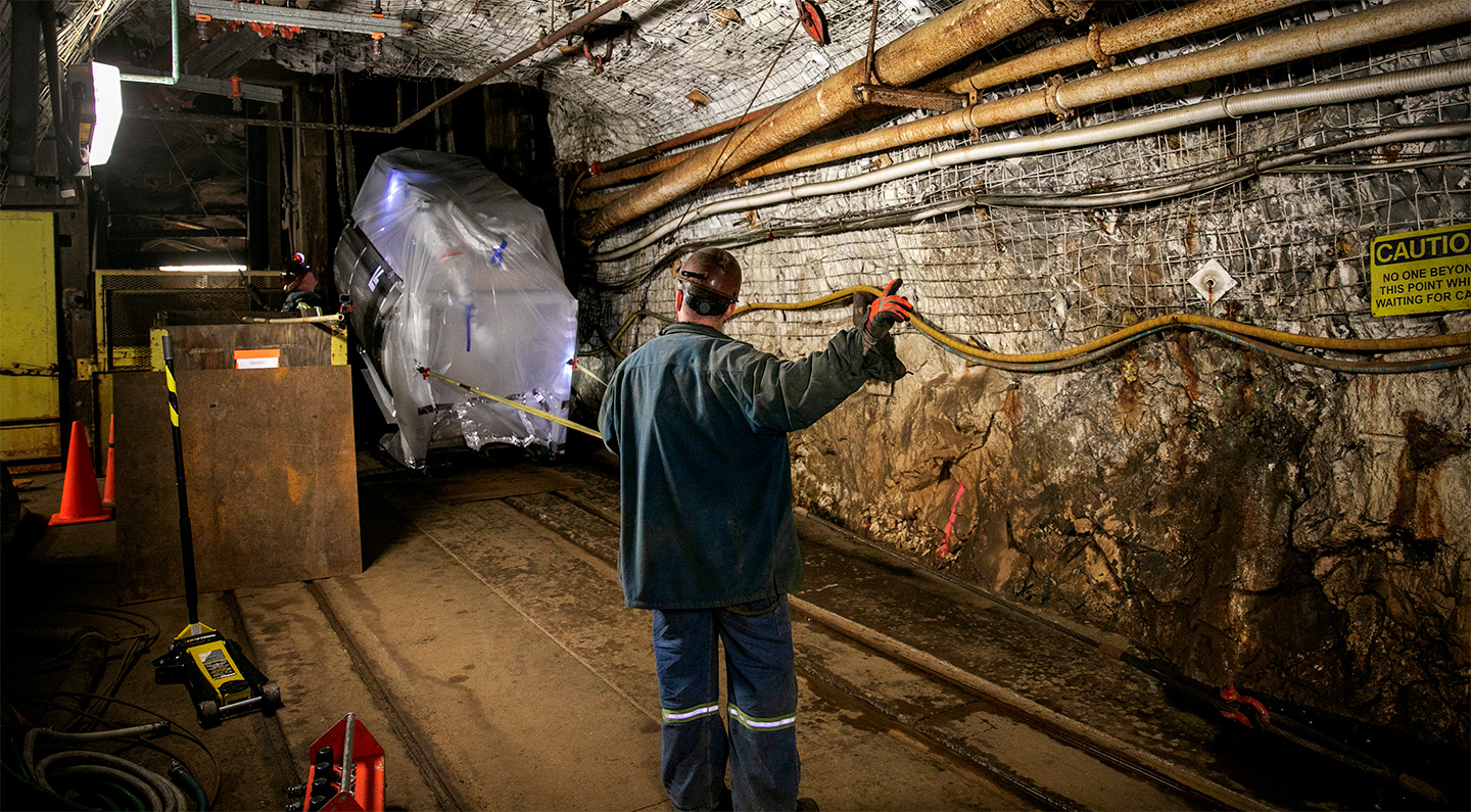 A worker in protective gear maneuvers a large, covered object within a dimly lit underground tunnel. The environment features exposed rocks, pipes, and caution signs, highlighting safety protocols.