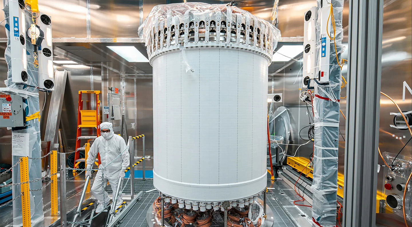 A researcher in protective gear works near a tall, cylindrical scientific instrument with visible wiring. The walls surrounding it are reflective metal.