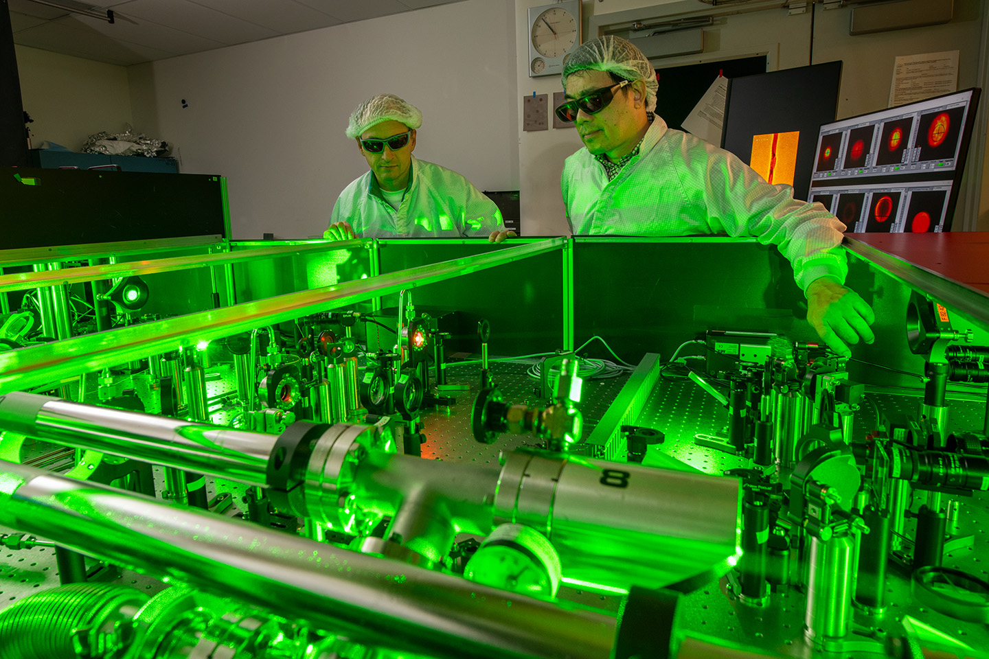 Two researchers in lab coats and safety glasses examine an optical table filled with metal components, illuminated by green light.