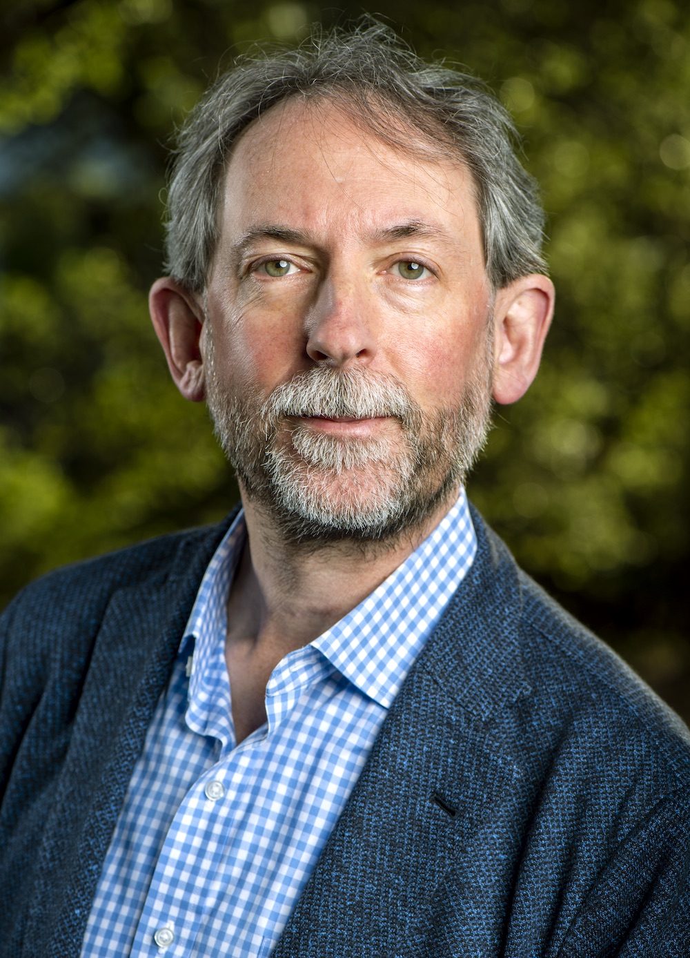 A man in a dark blue suit poses for a profile photo against a blurred green background